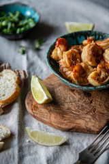 Turquoise bowl with steaming roasted garlic shrimps, lemon quarters, fresh baguette and bowl with greenery on linen tablecloths.