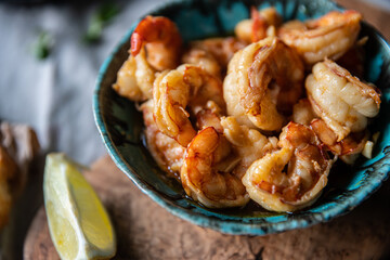 Turquoise bowl with steaming roasted garlic shrimps, lemon quarters, fresh baguette on linen tablecloths.
