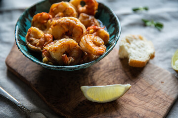 Turquoise bowl with steaming roasted garlic shrimps, lemon quarters, fresh baguette and bowl with greenery on linen tablecloths.