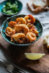 Turquoise bowl with steaming roasted garlic shrimps, lemon quarters, fresh baguette and bowl with greenery on linen tablecloths.