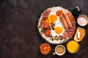 English traditional breakfast on a dark brown rustic background. Top view, flat lay, copy space. Fried eggs with bacon, sausages and beans, coffee, orange juice.
