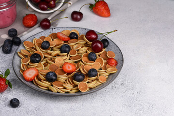 Tiny pancake cereal mini pancakes in a grey bowl with strawberries, blueberries sweet cherry on light background. Copy space