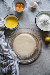 Step by step process of making yeast bread. Top view of ingredients: bowls with yeast starter, melted butter, egg yolks and flour, lemon, egg shells and towel on linen tablecloth.