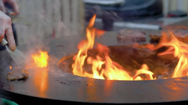 Close up view: chef grilling fresh meat steaks on brazier with hot flame at summer local food market - slow motion. Outdoor cooking, barbecue, gastronomy, cookery, street food concept