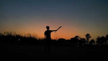 Silhouette of a boy playing cricket in the evening with the crescent moon in the beautiful sky full of variety of colors after sunset as wallpaper background. Gully cricket is very popular in India. © Dhananjay