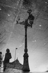 Reflection of an old-fashioned street lamp and a person in a pool of rainwater on a market square depicted in black and white.