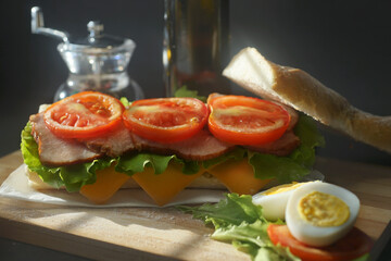 An appetizing sandwich with ham, cheese, tomatoes and lettuce lies on a board with egg and lemon  on black background