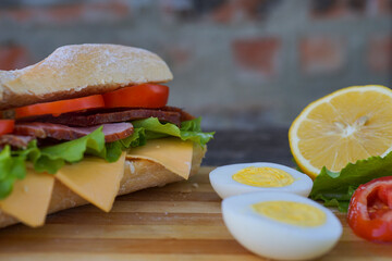 An appetizing sandwich with ham, cheese, tomatoes and lettuce lies on a board with egg and lemon