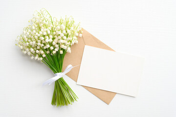 Bouquet of lilies of the valley and envelope with greeting card mockup on white table. Top view. Wedding, Mother's Day or Women's Day celebration concept.