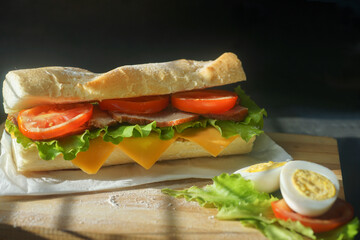 An appetizing sandwich with ham, cheese, tomatoes and lettuce lies on a board with egg and lemon  on black background