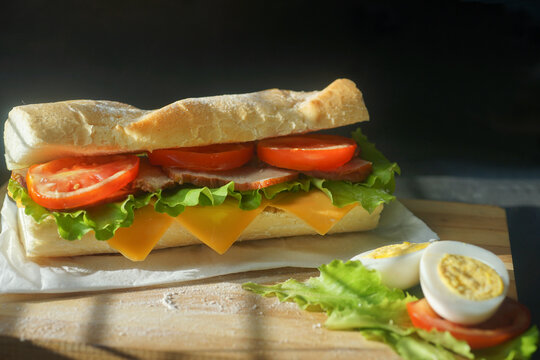 An Appetizing Sandwich With Ham, Cheese, Tomatoes And Lettuce Lies On A Board With Egg And Lemon  On Black Background