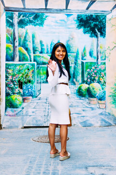 Latin brunette woman turned smiling and waving in front of street garden mural painted on wall.