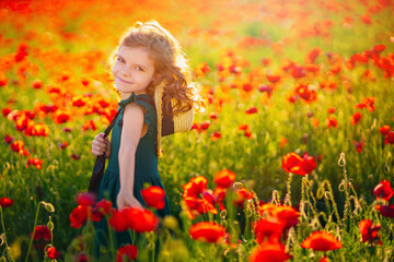 Naklejka na ściany i meble Cute little girl in green dress and straw hat posing at field of poppies on summer sunset.