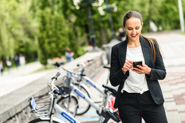 Fototapeta na wymiar Use environmentally friendly modes of transport. Young woman is using app on the phone for renting a city bike, she stands with a smartphone near parking of bicycle