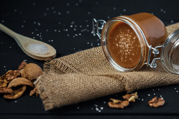 Glass jar of salted caramel with a wooden spoon of salt, walnuts and nutshells on a black wooden...