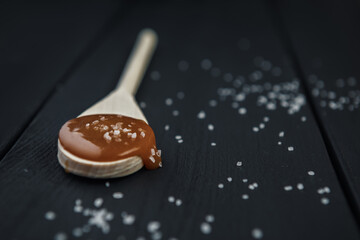 Wooden spoon with homemade salted caramel and crystals of sea salt on black wooden table.