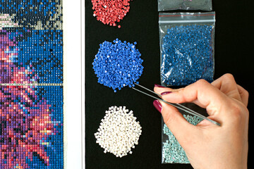 Girl collect diamond painting. Diamond embroidery with a tweezers. Acrylic rhinestones. Closeup,...