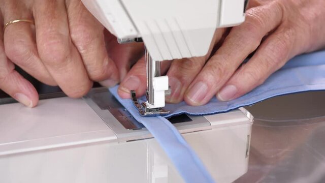 Elderly woman using hands to sew a blue cotton face mask with a modern sewing machine. Homemade reusable facial masks.