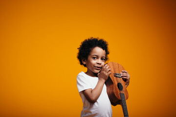 African American boy holding an inverted ukulele in his hands by his face and smiling playfully at camera on an orange background