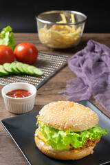 Crispy fried chicken burger on black plate served with french fries and ketchup on wooden background.