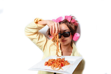 Curly girl in clothes and a plate with pasta and a fork wildly eats pasta with ketchup, standing in glasses on a white background. Girl eating spaghetti. Girl holding a plate of pasta.