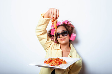 Curly girl in clothes and a plate with pasta and a fork wildly eats pasta with ketchup, standing in glasses on a white background. Girl eating spaghetti. Girl holding a plate of pasta.