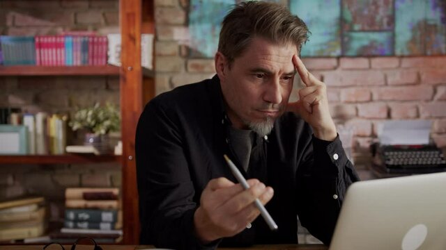 Serious older man working at home with laptop computer sitting at desk in home office thinking and waiting.