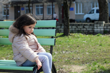 Little white cute smiling girl sits on a bench against the background of a blurry building cross-legged