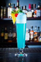 Blue drink cocktail with foam decorated pineapple and cherry in big glass, close up at the bar counter