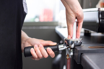 Fototapeta na wymiar close-up photo of female hands holding a metal tamper and a portafilter with coffee in a coffee shop. A barista preparing for pressing ground coffee for brewing espresso or americano in a cafe