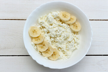Healthy breakfast. Cottage cheese in white bowl with banana on wood background