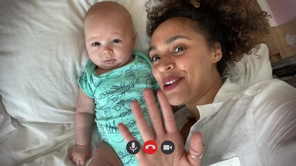 Mother and her newborn baby in bed making video call to family member using laptop on kitchen table - Powered by Adobe
