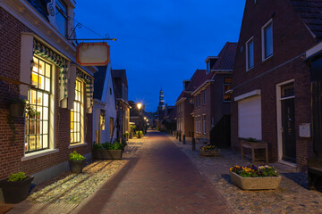 The Netherlands - Hindeloopen - A night alley lit by lamps and light from large shop windows. Typical Dutch architecture on both sides of the family houses. At the end of the alley is a church.