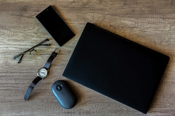 On a textured brown wooden table is a home work space with a black laptop, mobile phone, wristwatch, glasses, and computer mouse. Close-up view from above.