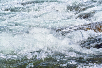 Rapid spring river flowing over rocks on sunny day, forming white water waves, closeup detail - abstract nature background