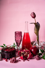 Still life in shades of red: glass bowl with cranberries, pomegranates, wine glass, bottle with beverage and tulips on pink background.