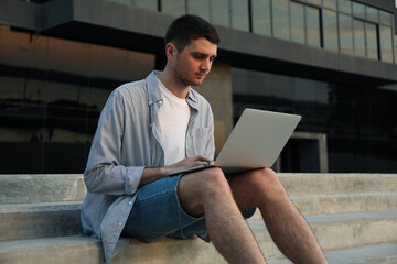 Young man sits in steps and working in laptop outdoor. Freelancer