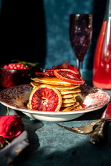 Vintage plate with pancakes with blood orange slices, red sauce and pomegranate seeds with sparkling glass goblet and bowl with cranberries  on dark background.