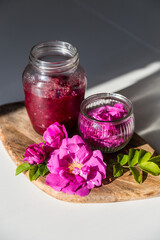 Glass jars with natural homemade rose jam and edible rose flowers on wooden board on white background.
