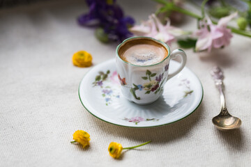 Espresso coffee in small delicate vintage cup with floral print, spoon and spring flowers on linen background.