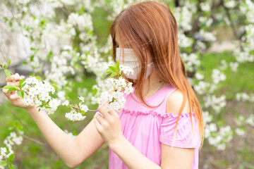 a girl in protective face mask stands near blossoming cherry tree during quarantine due to the coronavirus pandemic