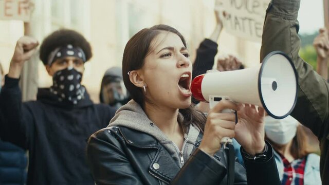 Pretty young Caucasian girl screaming in megaphone while standing among people at protest against racism and police brutality. Mixed-races male and female protesters.