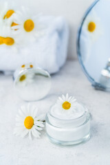 Herbal cosmetic cream in opened glass container with fresh chamomile flower, towel, mirror on a white background. Natural organic moisturizer skincare product. Selective focus. Vertical. Copy space.