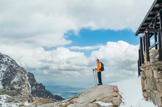 Dressed bright orange jacket backpacker walking in High Tatras mountain range, Slovakia using trekking poles. Hiker standing near Mountain hut at 2015m with kate snow around on the June beginning.