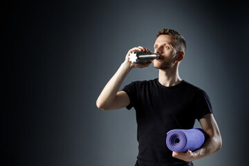 Fototapeta na wymiar Young man on the black background is drinking water from fitness bottle, holding fitness mat.