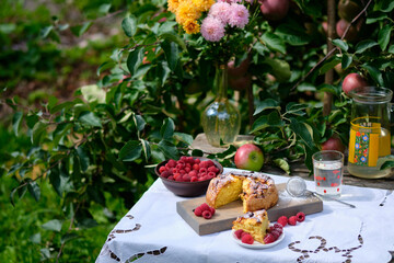 A piece cut from an apple pie lies on a plate. Rustic still life: sunny bright day, apple pie under an apple tree on an old wooden table, fruits and berries, flowers.