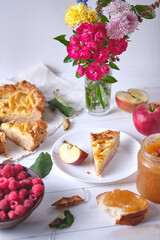 A sliced rustic apple pie on a linen towel. Dessert on a white wooden table, around are apples, raspberries in a plate, apple jam and flowers.