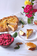 A sliced rustic apple pie on a linen towel. Dessert on a white wooden table, around are apples, raspberries in a plate, apple jam and flowers.