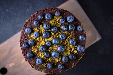 Chocolate cake with blueberries and dried cherries decorated with ground pistachios and berries on a wooden board.