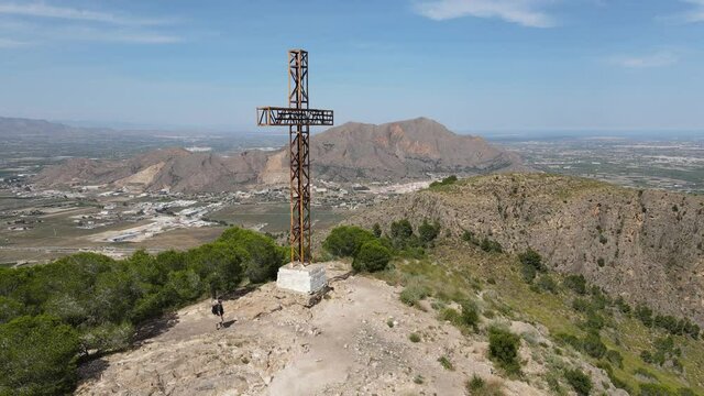 Mount Muela near the city of Orihuela. A hiker with a backpack reaches the summit cross and looks into the landscape with the drone standing still in the air. Sunshine and blue sky.